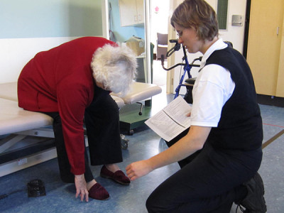 An elderly woman leans down from a hospital bed being assisted with her shoes by a physiotherapist 