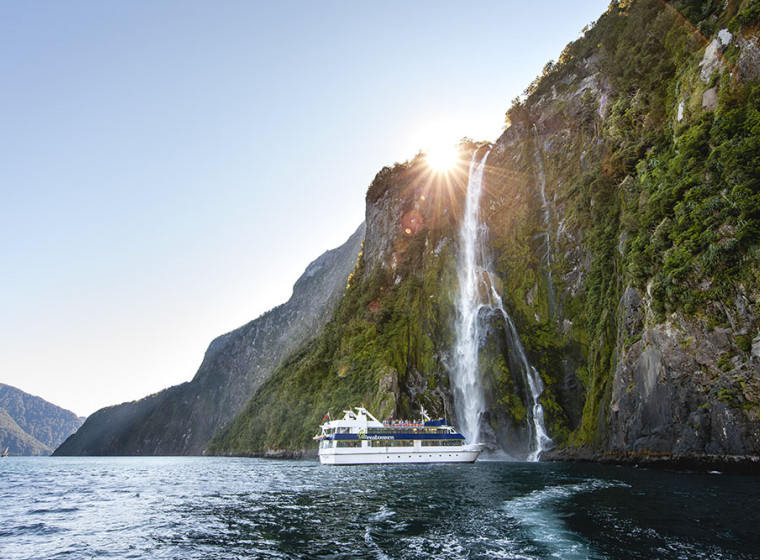 A boat on a lake with a waterfall behind it. The sun is setting behind the cliff face that the waterfall runs down
