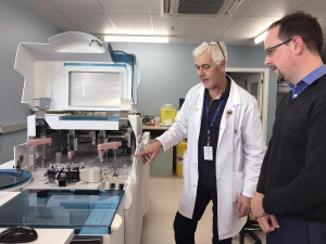 Two men, one in a lab coat, look at medical testing equipment 