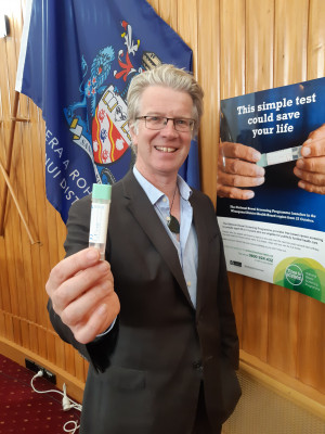 Whanganui Mayor Hamish McDouall holds a bowel screening test against the wood paneled walls of the Whanganui Council Chamber next to a bowel screening poster