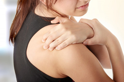 A young white woman with dark brown hair in a tank top massages the top of her shoulder