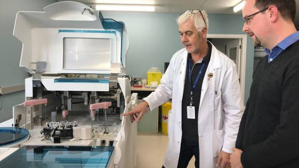 A man in a lab coat shows another man laboratory equipment  